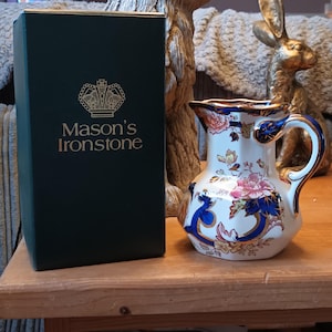 May include: A white ceramic jug with a blue and gold floral design. The jug is part of a Mason's Ironstone set. The jug is sitting on a wooden table.
