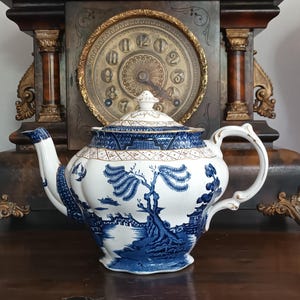 Puede incluir: Una tetera de porcelana blanca y azul con un diseño de paisaje. La tetera tiene un asa curva, un pico y una tapa con un adorno. El diseño incluye árboles, edificios y elementos acuáticos. La tetera está sobre una superficie de madera.