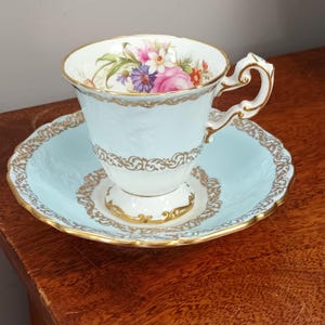 Op de afbeelding: Een delicaat theekopje en schotelset met een lichtblauwe basis en gouden rand. Het theekopje heeft een bloemenmotief met roze, gele en paarse bloemen.