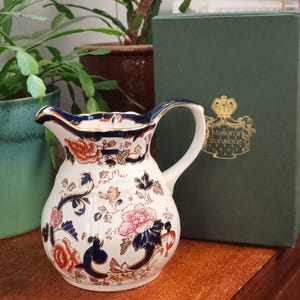 Puede incluir: Una jarra de cerámica blanca con un patrón floral azul y dorado. La jarra tiene una boca ancha y un asa. La jarra está sentada sobre una superficie de madera. El texto "Mason's Ironstone" es visible en la jarra.