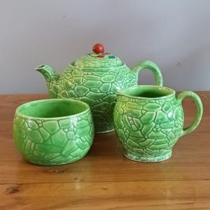 Op de afbeelding: Een groene keramische theeservies met een textuur, reptielachtig patroon. De set bevat een theepot met een rode knop, een melkkan en een kleine kom. De theeset wordt gepresenteerd op een houten oppervlak.