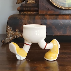 Puede incluir: Un divertido huevero de cerámica blanquecina con piernas y pies. Las piernas son blancas con una raya roja, y los pies son amarillos con detalles blancos. El huevero está sobre una superficie de madera oscura.