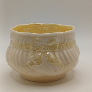 May include: A small, white ceramic bowl with a yellow glaze. The bowl features a raised design of a bow on the front.