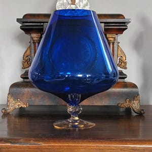 Peut inclure: Un grand vase en verre bleu cobalt, avec une base large et arrondie et un sommet évasé. Le vase repose sur un pied clair et travaillé avec un design torsadé. Il est présenté sur une surface en bois foncé, avec des éléments décoratifs ornés en arrière-plan.