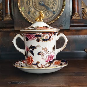 Puede incluir: Un azucarero de porcelana blanca con dos asas y una tapa a juego con un remate dorado en forma de animal. El cuenco y el plato están decorados con un patrón floral en tonos rosa, naranja y azul oscuro, con detalles en borde dorado.