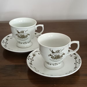 Pode incluir: Duas chávenas de chá brancas e pires com um design floral dourado. As chávenas apresentam um logotipo para Hendrick's Gin, um "A Most Unusual Gin" com um gráfico de uma ilustração botânica.