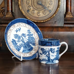 May include: A blue and white porcelain teacup and saucer set with a landscape design. The cup and saucer have a gold trim. The set is displayed in front of a vintage clock.