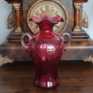 May include: A deep red glass vase featuring a ruffled rim and two clear glass handles on the sides. A small, partially visible sticker on the front of the vase contains the text "Fenton" and "Authentic Glass" with a small logo above the lettering.