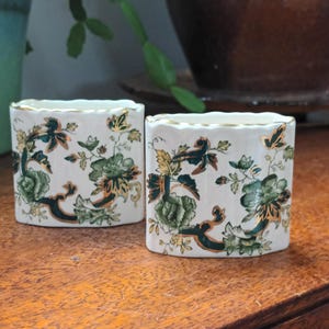 Może przedstawiać: Dwa małe, białe wazony ceramiczne z zielonym i złotym wzorem kwiatowym. Wazony mają owalny kształt i falisty brzeg.