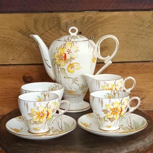 Puede incluir: Un juego de té vintage con tetera, jarra de leche, azucarero y dos tazas con platillos. El juego es blanco con un diseño floral en amarillo, naranja y verde. El juego se exhibe sobre una superficie de madera.