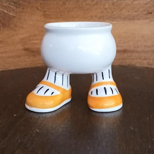 Puede incluir: Una maceta de cerámica blanca caprichosa con un cuerpo redondeado y patas. Las patas están pintadas con calcetines a rayas blancas y negras y zapatos naranjas con detalles negros. La maceta está diseñada para colocarse sobre una superficie plana.