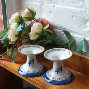 Puede incluir: Dos candelabros de cerámica blanca con diseños decorativos azules y marrones. Los candelabros tienen una base de pedestal y una parte superior circular. El diseño incluye un motivo de pájaro azul. Los candelabros se exhiben en una estantería de madera.