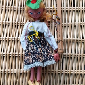 Puede incluir: Una muñeca de marioneta Pelham vintage con cabello rubio, ojos azules y un sombrero verde con una flor roja. La muñeca lleva una blusa blanca, una falda floral negra y zapatos de madera. La marioneta está sobre un fondo tejido.