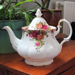 Puede incluir: Una tetera de porcelana blanca con un patrón floral de rosas y hojas. La tetera tiene un borde dorado y una tapa con un pomo en forma de rosa.