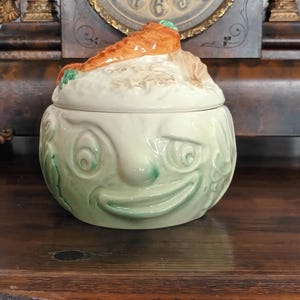 May include: A whimsical ceramic jar with a lid, featuring a face design with a carrot on top. The jar is primarily off-white with green and orange accents. The jar has a smiling face design.