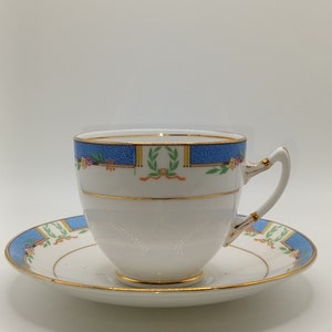 May include: White teacup and saucer set with a blue and gold decorative border. The cup and saucer feature a floral and ribbon design. The rim and handle are trimmed with gold. A classic tea set.