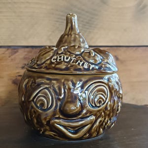 May include: Brown ceramic chutney jar with a smiling face design. The lid is topped with a small, textured knob and features the word "CHUTNEY". The jar has a round body with a textured surface.