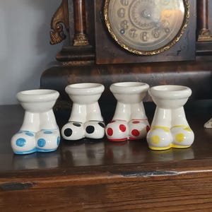 Puede incluir: Cuatro candelabros de cerámica con forma de piernas, cada uno con una parte superior blanca y partes inferiores de lunares de colores o de color sólido. Los colores incluyen azul, negro, rojo y amarillo. Los candelabros se exhiben sobre una superficie de madera.