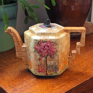 Może przedstawiać: Zabytkowy, geometryczny czajnik z jasnożółtym wzorem ceglanym. Czajnik posiada kwiatowy wzór z różowymi kwiatami i brązowym drzewem. Pokrywka ma mały czarny uchwyt. Czajnik stoi na drewnianej powierzchni.