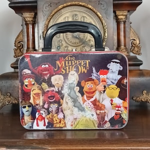 Può includere: Un portapranzo rettangolare in metallo con una grafica colorata di vari personaggi dei Muppet e la scritta The Muppet Show. Il portapranzo ha una maniglia in plastica nera sulla parte superiore e mostra segni di usura e perdita di vernice.