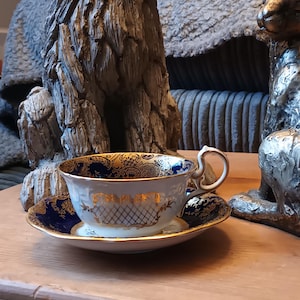 May include: A white teacup and saucer with a blue and gold floral pattern. The cup has a gold rim and a gold handle. The saucer has a blue rim and a gold floral pattern.