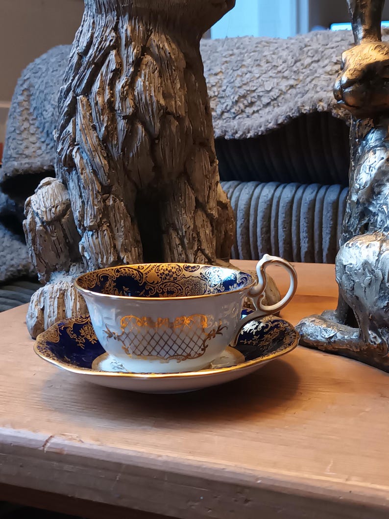 May include: A white teacup and saucer with a blue and gold floral pattern. The cup has a gold rim and a gold handle. The saucer has a blue rim and a gold floral pattern.