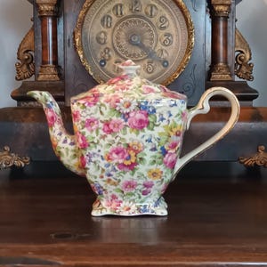 Pode incluir: Uma chaleira vintage com um padrão floral em rosa, amarelo e azul. Possui um bico curvo e uma alça com acabamento dourado. A chaleira está sobre uma superfície de madeira escura, com um relógio ao fundo.