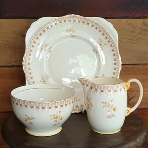 Può includere: Un set di piatti vintage color crema con un piatto quadrato, una ciotola e una brocca. Ogni pezzo è decorato con un delicato motivo floreale e un bordo decorativo nei toni del marrone e del giallo. Il set è esposto su una superficie di legno.