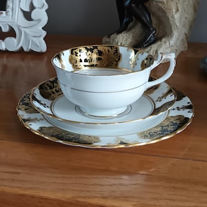 May include: A white teacup and saucer set with black and gold floral designs. The cup has a gold rim and a gold band around the middle. The saucer has a scalloped edge.