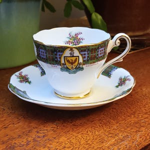 May include: A white teacup and saucer with a gold trim. The cup and saucer feature a green, blue, and red plaid pattern with a crest in the center. The crest features a golden eagle with a red and white shield. The text "Clan Macdonald" is printed on the cup and saucer.