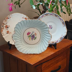 May include: Three decorative plates with floral designs. The plates are on a wooden table with a drawer. The plates are in different sizes and colors. The largest plate is light blue with a floral design in the center. The middle plate is white with a gold trim and a floral design. The smallest plate is white with a gold trim and a floral design.