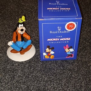 May include: A ceramic figurine of Goofy from Disney's Mickey Mouse. Goofy is wearing a blue shirt, brown trousers, and a green hat. He is sitting on a white base. The figurine is part of the Royal Doulton Mickey Mouse Collection.