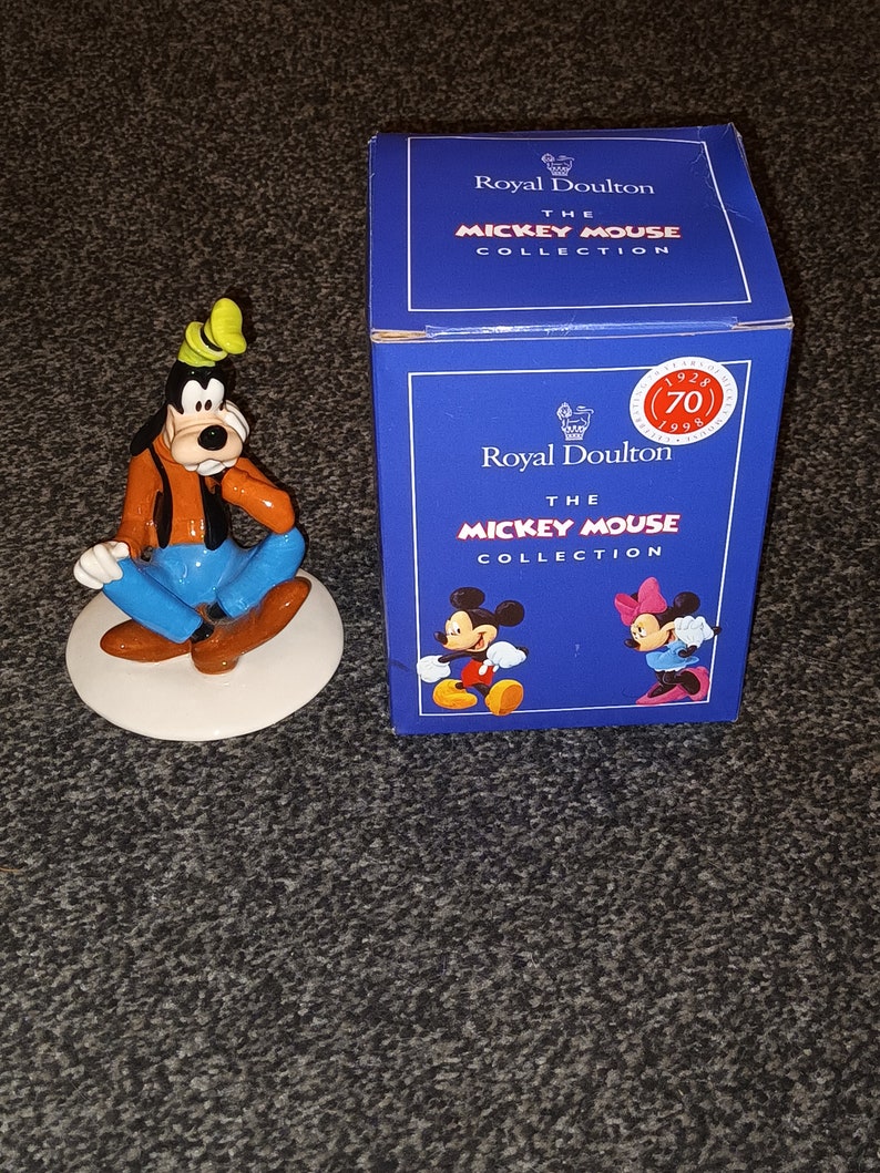 May include: A ceramic figurine of Goofy from Disney's Mickey Mouse. Goofy is wearing a blue shirt, brown trousers, and a green hat. He is sitting on a white base. The figurine is part of the Royal Doulton Mickey Mouse Collection.