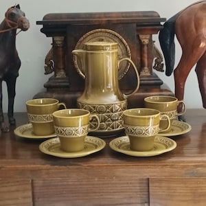 Può includere: Un set da caffè vintage in ceramica verde oliva composto da una caraffa con coperchio e quattro tazze abbinate con piattini. Ogni pezzo presenta una fascia geometrica strutturata con motivi circolari ed è rifinito con uno smalto lucido.