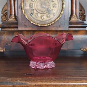 May include: A deep magenta glass bowl featuring a wide, ruffled rim and a textured, clear glass base. The bowl has a smooth, translucent finish and a curved, open design. It is positioned in front of a dark wooden clock with a gold-toned face.