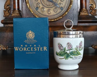 May include: A white ceramic egg cup with a silver lid and handle, decorated with a botanical design of green leaves and purple flowers. A blue box with gold lettering that reads "ROYAL WORCESTER" is next to the egg cup.