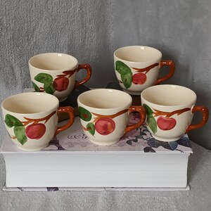 Pode incluir: Conjunto de cinco chávenas de chá de cerâmica cor de creme com alças marrons. Cada chávena apresenta um desenho de maçã vermelha e folhas verdes num ramo castanho.