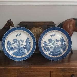 May include: Two round ceramic plates featuring a blue and white willow pattern. Each plate has a scalloped edge and a central illustration of a bridge, a large tree, and two birds in flight. The design includes intricate geometric borders.