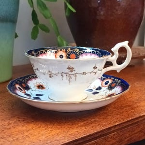 以下が含まれることがあります： 青と金の色の花柄が描かれた白いティーカップとソーサー。カップは縁がギザギザになっていて、持ち手があります。ソーサーはカップと同じ柄です。