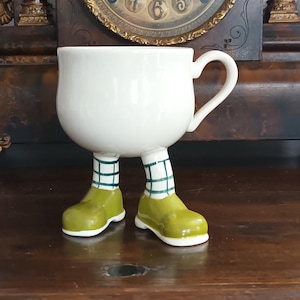 Könnte beinhalten: Eine skurrile weiße Keramik-Teetasse mit Henkel, die auf zwei Beinen steht. Die Beine sind mit grünen Schuhen und weiß-grün karierten Socken versehen. Die Tasse steht auf einer Holzoberfläche.