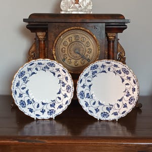 May include: Two white porcelain plates with a blue floral pattern and gold trim. The plates are on white stands in front of a vintage clock with a dark wooden frame. The clock has a gold-coloured face.