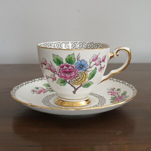 May include: A white teacup and saucer with a floral pattern in shades of pink, blue, yellow, and green. The cup and saucer have a gold trim.