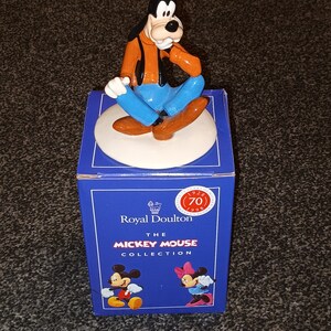 May include: A ceramic figurine of Goofy from Disney's Mickey Mouse. Goofy is sitting with his legs crossed and wearing a blue shirt, orange jacket, and a green hat. The figurine is on a white base and is part of the Royal Doulton Mickey Mouse Collection.