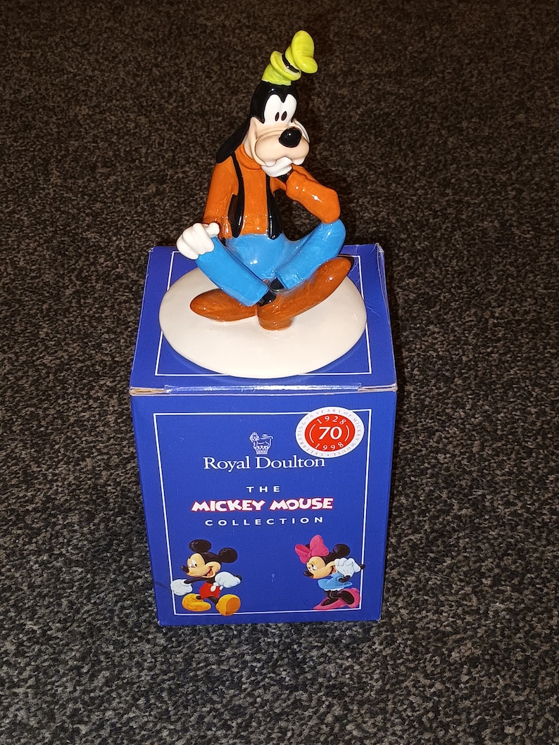May include: A ceramic figurine of Goofy from Disney's Mickey Mouse. Goofy is sitting with his legs crossed and wearing a blue shirt, orange jacket, and a green hat. The figurine is on a white base and is part of the Royal Doulton Mickey Mouse Collection.