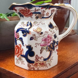 Może przedstawiać: Biały ceramiczny dzbanek o sześciokątnym kształcie, ozdobiony kolorowym wzorem kwiatowym w odcieniach czerwieni, różu i pomarańczu, podkreślony ciemnoniebieskimi zawijasami oraz złotym wykończeniem wzdłuż falistego brzegu i krawędzi uchwytu.