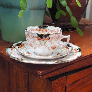 Puede incluir: Un juego de taza y platillo de té vintage con un patrón floral en tonos naranja, verde y negro. La taza tiene un borde festoneado y el platillo tiene un borde dorado.