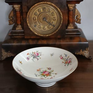 May include: A white porcelain bowl with a floral design in pink, yellow, and purple. The bowl has a gold rim and sits on a pedestal base. A vintage clock is in the background.