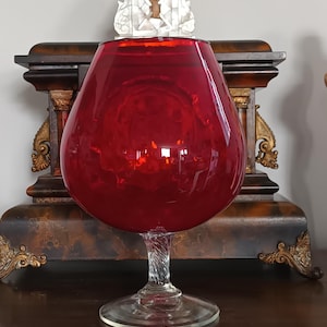 May include: A large, ruby-red glass brandy snifter. The bowl-shaped glass sits atop a clear, twisted stem and a circular base. The glass reflects light, creating a vibrant display. The snifter is a decorative piece, suitable for a home bar.