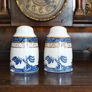 Op de afbeelding: Twee bijpassende witte keramische zout- en peperstrooiers met blauwe en gouden decoratieve banden. De strooiers hebben een blauw wilgenboomontwerp. De strooiers staan op een houten oppervlak.