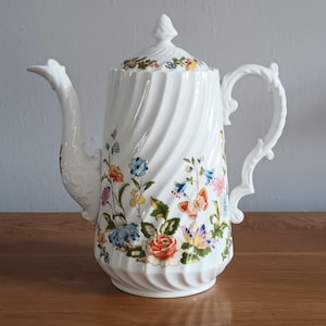 May include: White porcelain coffee pot with a lid, featuring a floral design with butterflies. The pot has a spiral design and a curved handle. The flowers are in shades of blue, orange, and yellow.
